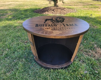 May include: A wooden barrel table with a black Buffalo Trace Kentucky Straight Bourbon Whiskey logo on the top. The table has a black interior and a metal band around the bottom.