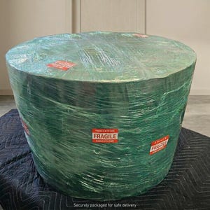 May include: A large, cylindrical object wrapped in green plastic. Red "FRAGILE" stickers are visible. The object is on a black quilted surface. The text "Securely packaged for safe delivery" is at the bottom.