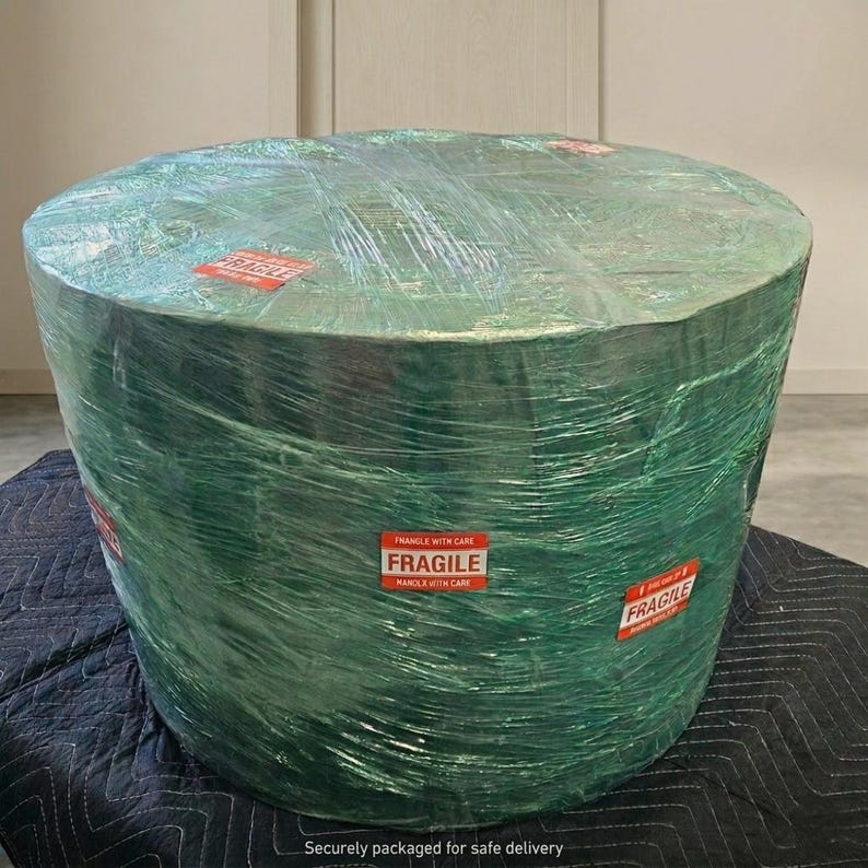May include: A large, cylindrical object wrapped in green plastic. Red "FRAGILE" stickers are visible. The object is on a black quilted surface. The text "Securely packaged for safe delivery" is at the bottom.