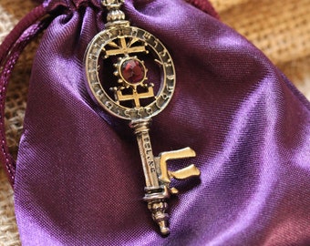 May include: A silver key pendant with a red gemstone in the center, surrounded by a circular design. The key is hanging from a purple satin pouch.