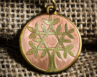 May include: A round pendant with a copper-colored center and a gold-colored border and bail. The pendant features a tree design with a Star of David at the center. The pendant is displayed on a textured, woven background.
