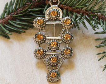 May include: A silver pendant necklace with a tree of life design. The pendant is adorned with multiple small, round, amber-colored rhinestones.