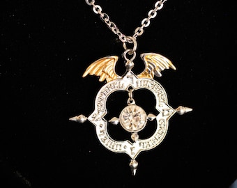 May include: A silver chain necklace with a pendant featuring a silver compass with a clear crystal in the center. The compass is surrounded by gold wings and has an inscription around the edge.
