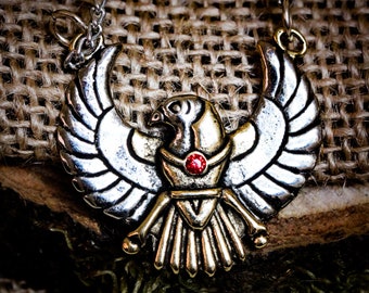 May include: A silver and gold pendant necklace featuring a stylized bird with outstretched wings. The bird has a red gemstone in its chest.