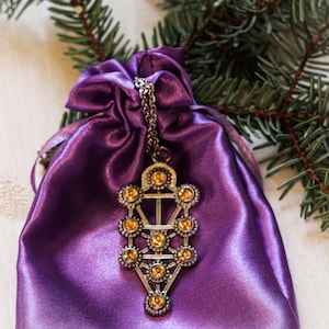 May include: A purple satin pouch with a silver pendant featuring a Tree of Life design with amber-colored crystals.