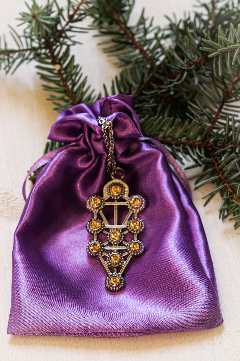 May include: A purple satin pouch with a silver pendant featuring a Tree of Life design with amber-colored crystals.