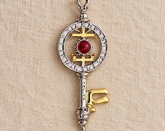 May include: A silver-toned metal pendant shaped like a key, featuring a circular head with a central red cabochon stone, gold-colored cross accents, and a textured outer ring. The shaft includes a gold-colored bit and a small loop at the top.
