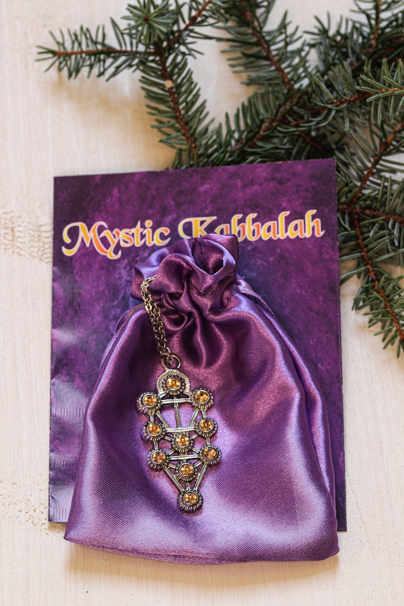 May include: A purple satin pouch with a silver Tree of Life pendant with amber-colored crystals. The pouch is on a card that says "Mystic Kabbalah".