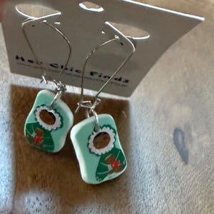 May include: A pair of dangle earrings with a light blue background and a painted design. The design features a brown-haired figure wearing a green coat with a white collar and a red accent. The earrings hang from silver-colored hooks.
