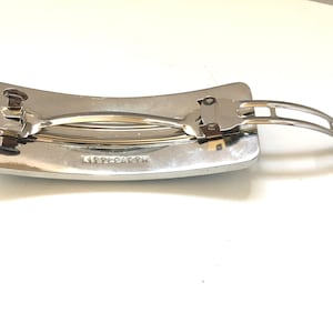 May include: A silver-toned hair barrette with a rectangular base and a curved top. The barrette features a hinged clasp and the text "LITTLE EARTH" is visible on the base.
