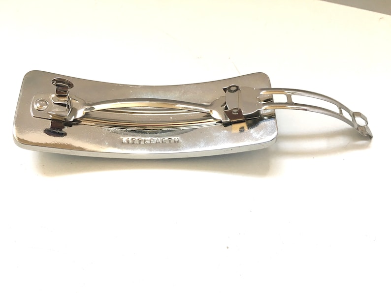 May include: A silver-toned hair barrette with a rectangular base and a curved top. The barrette features a hinged clasp and the text "LITTLE EARTH" is visible on the base.