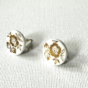 以下が含まれることがあります： 白いスタッドピアス2つ。金色の花の模様が施されています。ピアスは丸く、ポストバックが付いています。花の模様は細かく、ピアスにエレガントさを加えています。