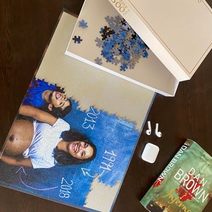 May include: A 500-piece jigsaw puzzle with a photo of a woman and child, with the years 1991, 2013, and 2018. The puzzle box is open, with loose pieces. A copy of Dan Brown's "Inferno" and a pair of wireless earbuds are also present.