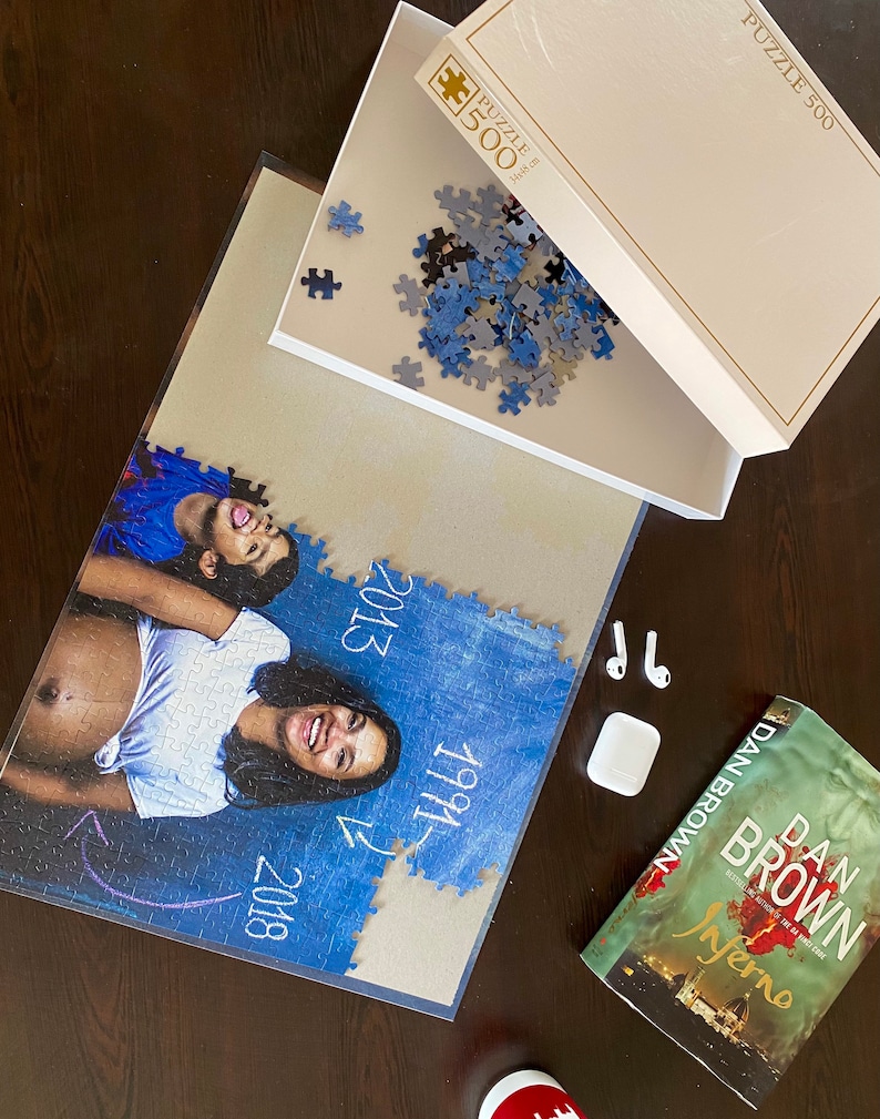 May include: A 500-piece jigsaw puzzle with a photo of a woman and child, with the years 1991, 2013, and 2018. The puzzle box is open, with loose pieces. A copy of Dan Brown's "Inferno" and a pair of wireless earbuds are also present.