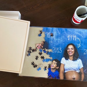 May include: A 500-piece family photo jigsaw puzzle, partly assembled. The image features a couple and a child, with the years 1991, 2013, and 2018 written on a blue backdrop. The puzzle box is off-white.