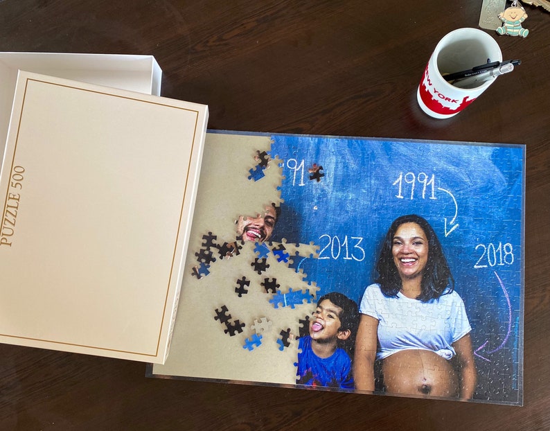 May include: A 500-piece family photo jigsaw puzzle, partly assembled. The image features a couple and a child, with the years 1991, 2013, and 2018 written on a blue backdrop. The puzzle box is off-white.