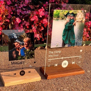 May include: Two clear acrylic photo frames with wooden bases. One frame displays a photo of two people, the other shows two people in graduation gowns. The text "MIXGIFT" is visible on both frames. The second frame has the text "The Climb" and "Miley Cyrus".