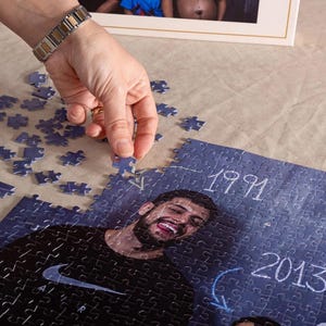 Puede incluir: Un rompecabezas fotográfico personalizado con un fondo azul, que presenta a una persona sonriente y los años 1991 y 2013. Una mano está colocando una pieza del rompecabezas. El rompecabezas está sobre una superficie beige.