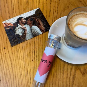 May include: A small jigsaw puzzle showing a couple, a clear vial with a pink heart label reading "love you", and a glass of coffee with latte art, all on a wooden table.