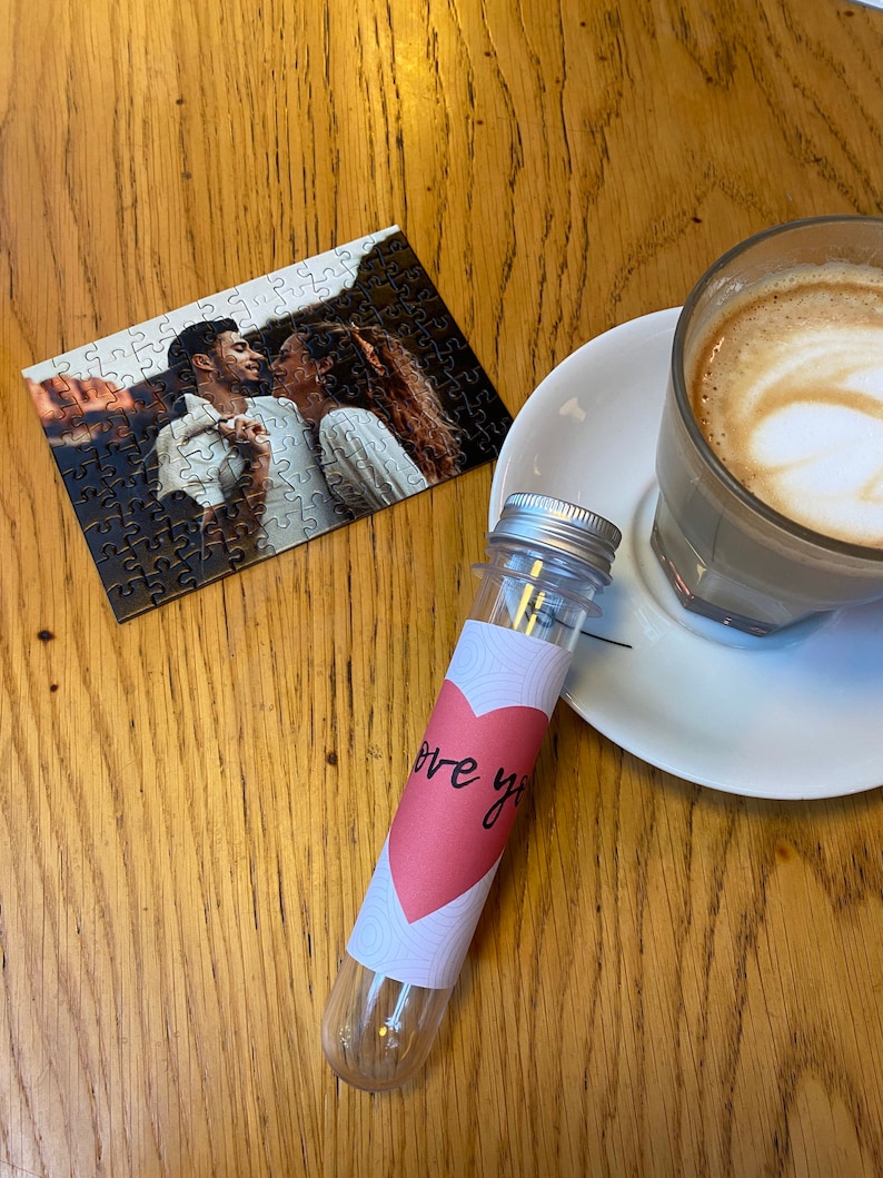 May include: A small jigsaw puzzle showing a couple, a clear vial with a pink heart label reading "love you", and a glass of coffee with latte art, all on a wooden table.