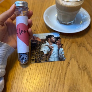 May include: A clear tube with a silver lid filled with puzzle pieces. The tube has a label with a pink heart and the words "I love". A partially assembled photo puzzle and a coffee cup are also visible.