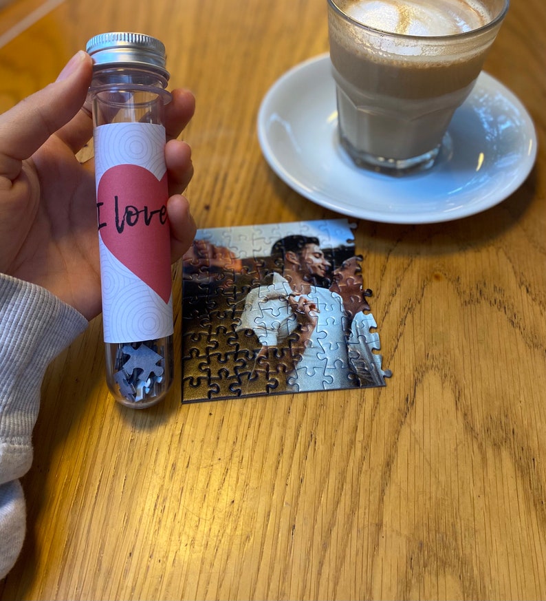 May include: A clear tube with a silver lid filled with puzzle pieces. The tube has a label with a pink heart and the words "I love". A partially assembled photo puzzle and a coffee cup are also visible.