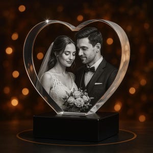 May include: A heart-shaped crystal photo frame displaying a wedding portrait of a couple. The frame is set on a black base. The couple is in formal attire, the bride holding a bouquet of roses. The background has blurred bokeh lights.