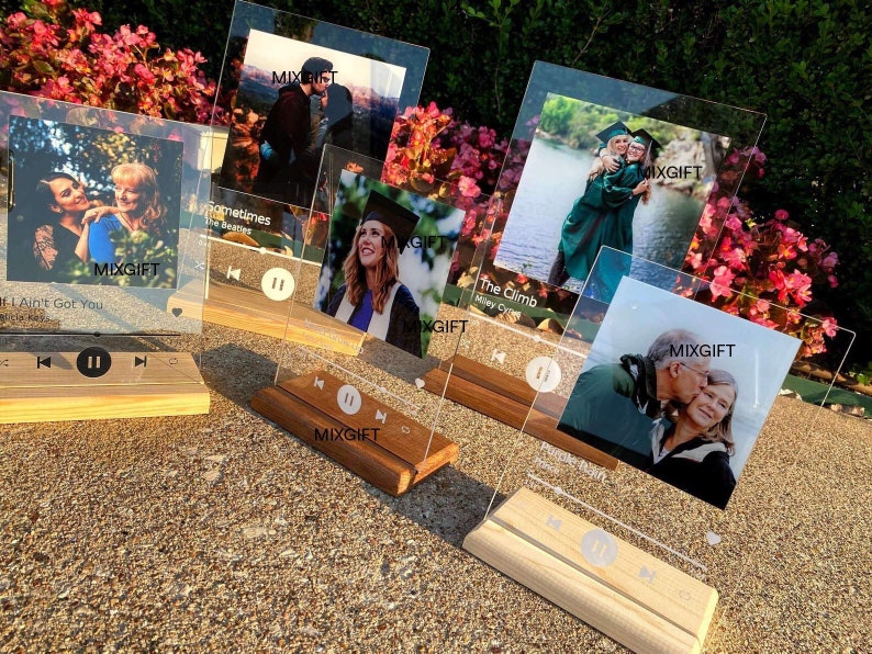 May include: Several clear acrylic photo displays with wooden bases are arranged outdoors. Each display features a printed photo, song title, and artist name, along with play/pause controls. The displays are personalized gifts.