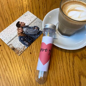 May include: A clear plastic tube with a silver lid and a pink heart-shaped label reading "love you". A small jigsaw puzzle featuring a photo of two people is next to the tube. A cup of coffee on a white saucer is also visible.