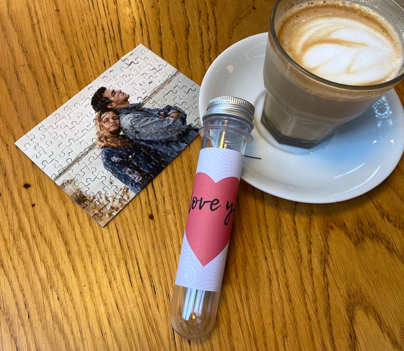 May include: A clear plastic tube with a silver lid and a pink heart-shaped label reading "love you". A small jigsaw puzzle featuring a photo of two people is next to the tube. A cup of coffee on a white saucer is also visible.