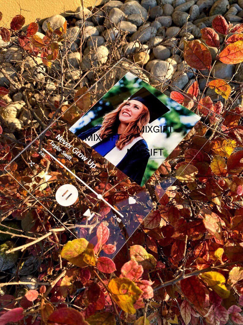 May include: A clear acrylic plaque featuring a photo of a woman in a graduation cap and gown. The plaque has a music player interface design with the song title "Never Grow Up" by Taylor Swift. The background includes colorful autumn leaves.