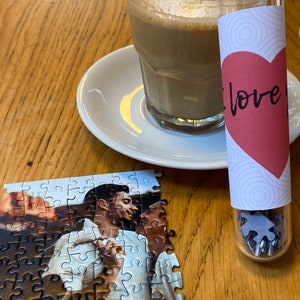 May include: A partially assembled jigsaw puzzle featuring a couple, with a glass of coffee and a small tube containing puzzle pieces. The tube has a red heart with the word "love" on it. The items are on a wooden table.