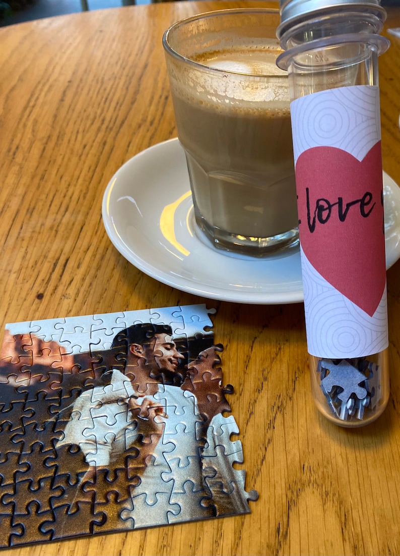 May include: A partially assembled jigsaw puzzle featuring a couple, with a glass of coffee and a small tube containing puzzle pieces. The tube has a red heart with the word "love" on it. The items are on a wooden table.
