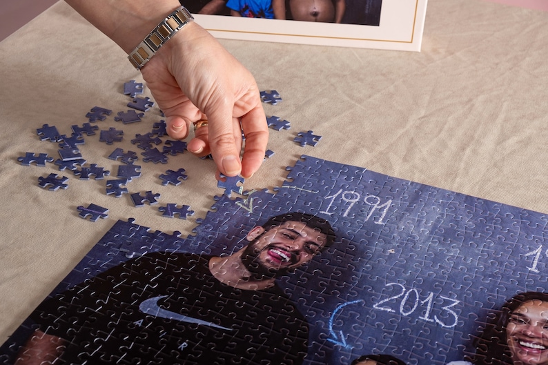 May include: A personalised photo jigsaw puzzle featuring a smiling person, the years 1991 and 2013. A hand is placing a puzzle piece. The puzzle is on a beige surface, with scattered pieces nearby. A framed photo is in the background.