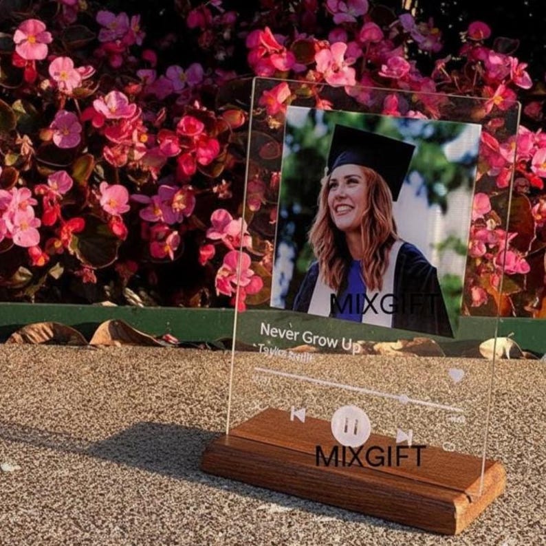 May include: A clear acrylic photo display with a wooden base. The display features a photo of a smiling person in a graduation cap and gown. The text "Never Grow Up" and "MIXGIFT" are visible.