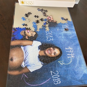May include: A 500-piece jigsaw puzzle featuring a family portrait. The image shows a pregnant person and a child, with the years 1991, 2013, and 2018 written on the puzzle. The puzzle box is white.