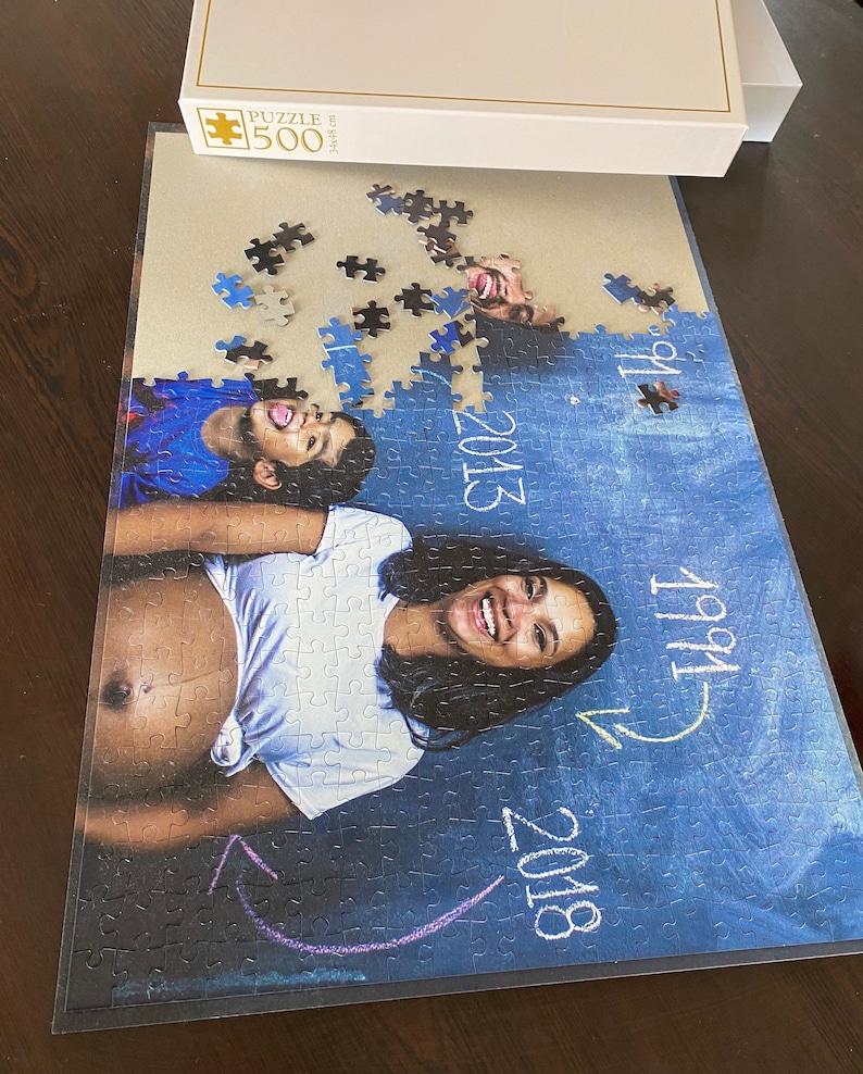 May include: A 500-piece jigsaw puzzle featuring a family portrait. The image shows a pregnant person and a child, with the years 1991, 2013, and 2018 written on the puzzle. The puzzle box is white.