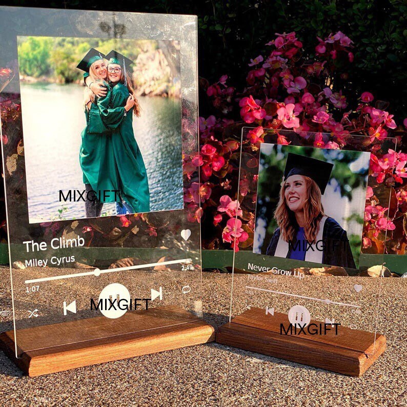 May include: Two acrylic music player-styled photo frames display graduation photos. The larger frame shows two graduates embracing, with 'The Climb' by Miley Cyrus displayed below. The smaller frame features a single graduate, with 'Never Grow Up' by Taylor Swift shown below. Both frames have wooden bases and a minimalist design.  The text 'MIXGIFT' is visible on both frames.