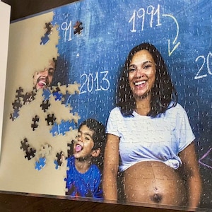 May include: A 500-piece jigsaw puzzle with a family portrait, including a pregnant woman. The image on the puzzle shows the years 1991, 2013, and 2018 on a blue background. The puzzle box is white and has the text "PUZZLE 500".