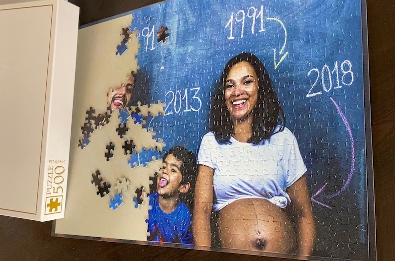 May include: A 500-piece jigsaw puzzle with a family portrait, including a pregnant woman. The image on the puzzle shows the years 1991, 2013, and 2018 on a blue background. The puzzle box is white and has the text "PUZZLE 500".