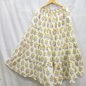 May include: A white tiered skirt featuring a repeating floral pattern in yellow and muted green. The skirt has an elasticized waistband with a drawstring and a full, flowing silhouette that falls in three distinct horizontal gathered tiers.