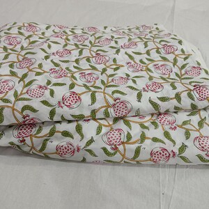 May include: A folded piece of white fabric featuring a repeating pattern of pink pomegranates with green leaves and yellow stems. The textile displays a botanical print with stylized fruit and vine motifs distributed evenly across the surface.