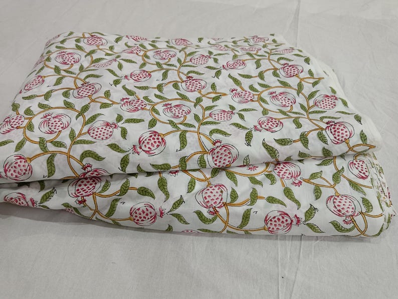 May include: A folded piece of white fabric featuring a repeating pattern of pink pomegranates with green leaves and yellow stems. The textile displays a botanical print with stylized fruit and vine motifs distributed evenly across the surface.