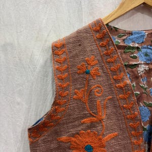 K&ouml;nnte beinhalten: Eine braune Weste mit orangefarbener Blumenstickerei und blauem Blumenmusterfutter. Die Weste h&auml;ngt an einem Holzb&uuml;gel. Die Stickerei zeigt eine gro&szlig;e Blume mit einem t&uuml;rkisfarbenen Zentrum.
