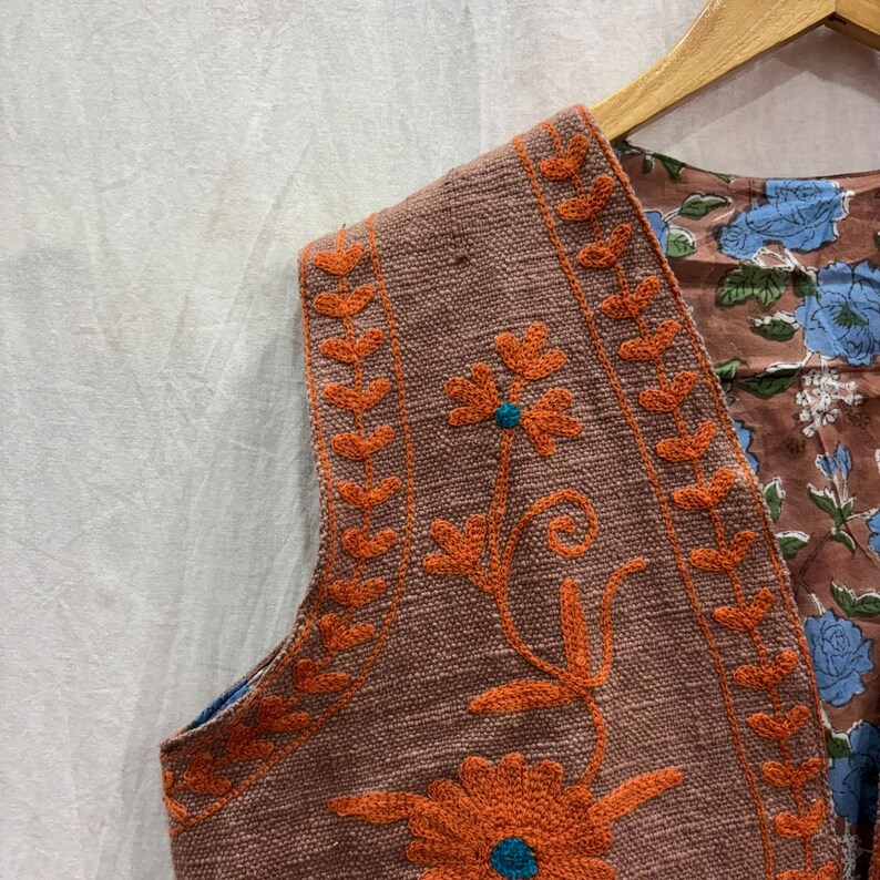 K&ouml;nnte beinhalten: Eine braune Weste mit orangefarbener Blumenstickerei und blauem Blumenmusterfutter. Die Weste h&auml;ngt an einem Holzb&uuml;gel. Die Stickerei zeigt eine gro&szlig;e Blume mit einem t&uuml;rkisfarbenen Zentrum.