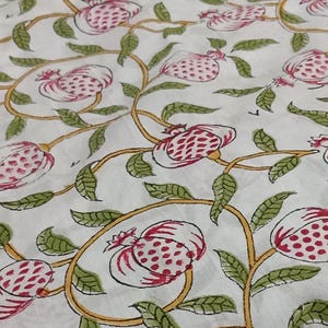 May include: A white fabric featuring a repeating pattern of pink pomegranates with green leaves and yellow vines. The design is printed across the surface, showcasing stylized fruit shapes and foliage connected by thin, flowing, curved yellow stems.