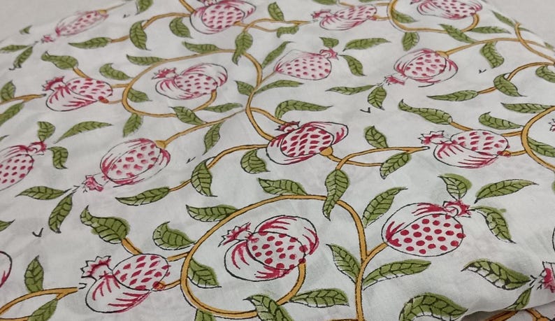 May include: A white fabric featuring a repeating pattern of pink pomegranates with green leaves and yellow vines. The design is printed across the surface, showcasing stylized fruit shapes and foliage connected by thin, flowing, curved yellow stems.