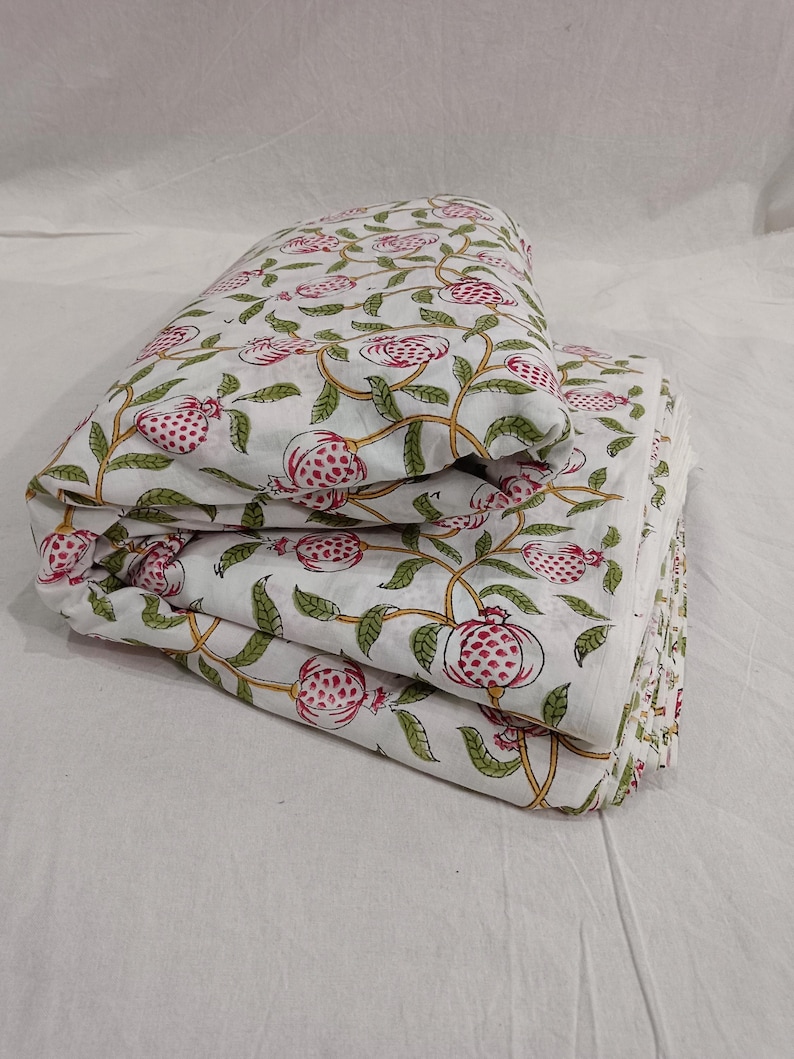 May include: Folded stack of white cotton fabric with a pink and green pomegranate print. The fabric features a repeating pattern of pomegranates on vines.  The design is block printed, showcasing a traditional Indian textile style. The fabric is suitable for quilting, apparel, or home decor projects.