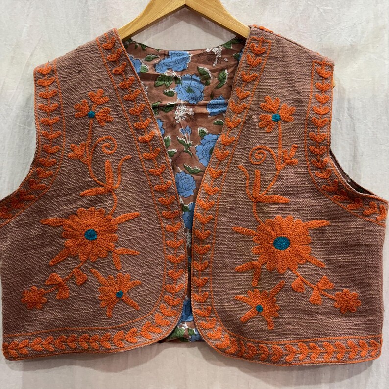 K&ouml;nnte beinhalten: Eine braune Weste mit orangefarbenen Blumenstickereien und einem blau gebl&uuml;mten Futter. Die &auml;rmellose Weste ist aus strukturiertem Stoff gefertigt. Die Stickerei zeigt Blumen mit t&uuml;rkisfarbenen Zentren.