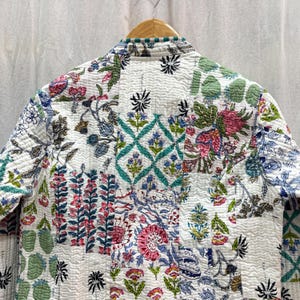 Pu&ograve; includere: Una giacca trapuntata bianca con un design a patchwork che presenta motivi floreali e geometrici in tonalit&agrave; di verde, blu, rosa e nero.